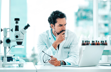 A scientist looking at laptop in a laboratory, with a microscope and various equipment in the background.
